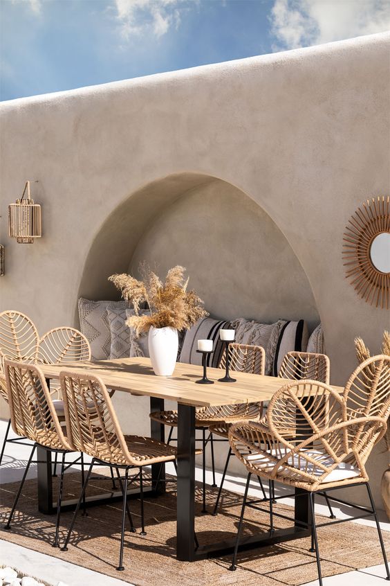 A modern boho outdoor dining space with a built in bench, a stained table, rattan furniture, a mirror and grasses in a vase