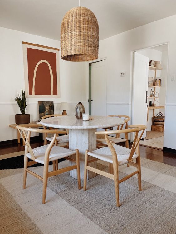 a modern dining room with layered rugs including a jute one, a round table, woven chairs, a woven pendant lamp, a bench with decor is a chic space