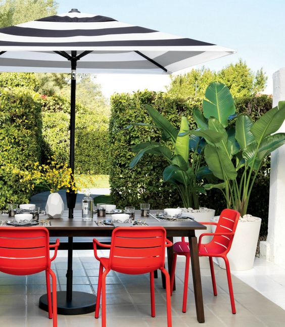 A modern dining space with a lot of greenery, a dark stained table, red chairs, a striped umbrella is a cool outdoor dining room