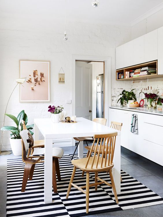 a modern eat-in kitchen with sleek white cabinets, a white table and mismatching chairs, a striped rug and some pretty decor