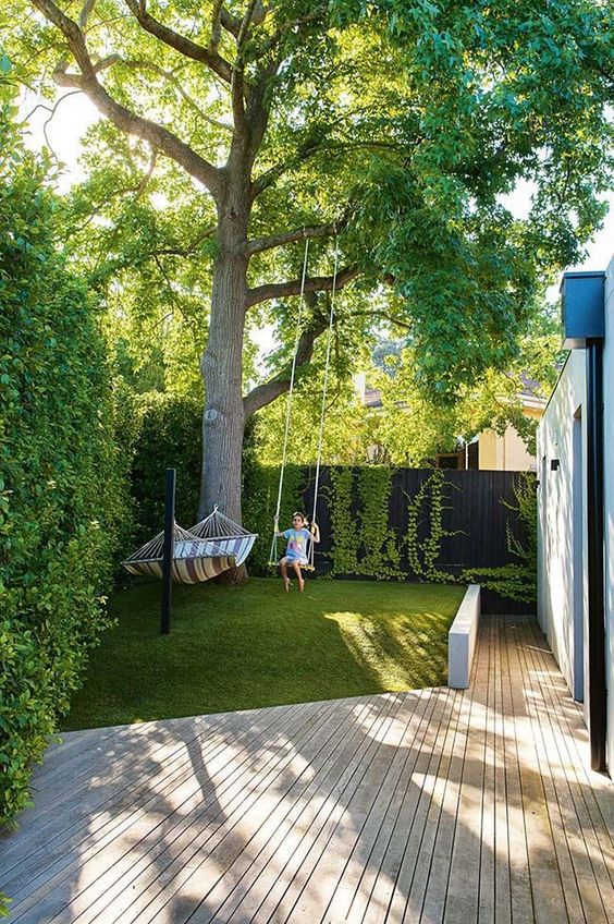 a modern garden with a deck, a green lawn, some trees and vines and a hammock plus a swing is amazing