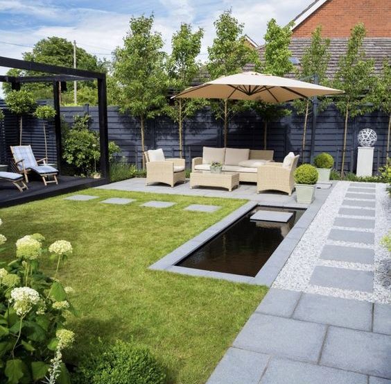 a modern garden with a green lawn, a black pergola with daybeds, a sitting area under an umbrella and some blooms