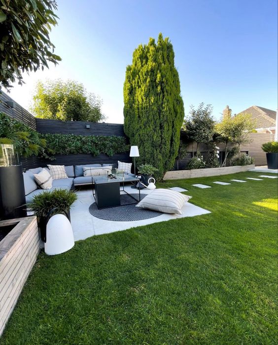 a modern garden with a green lawn, a living wall, some trees, a corner sofa, a coffee table and some pillows plus some lamps