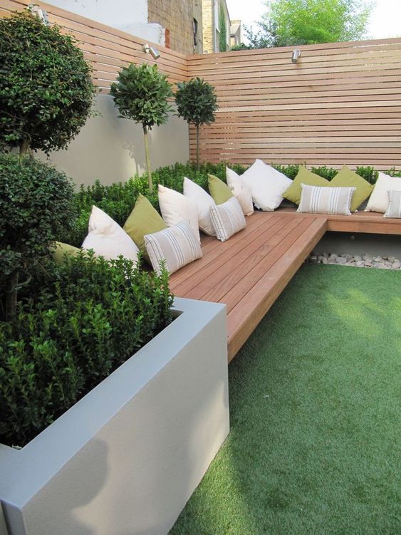 a modern garden with a green lawn, a raised garden bed with shrubs and trees, a built-in bench with some pillows