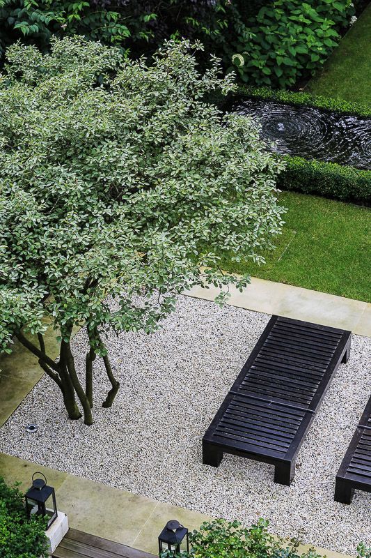 a modern garden with a green lawn, a water body, a tree, some pebbles and black wooden daybeds is cool