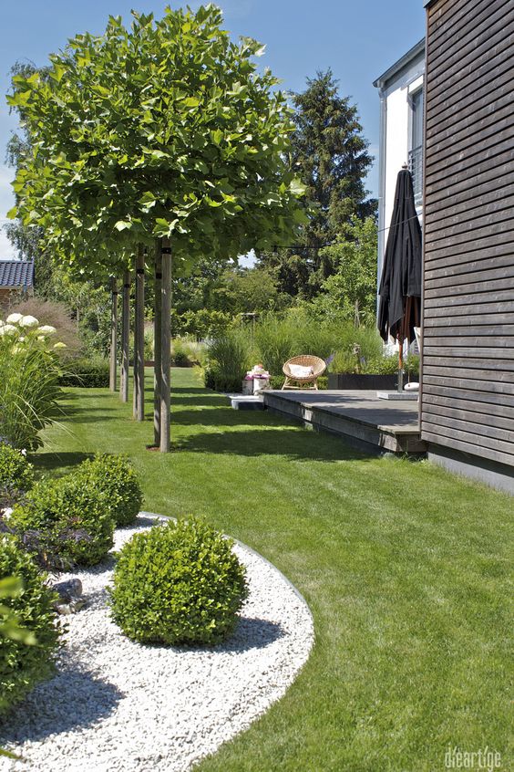 a modern garden with a green lawn and shrubs, some trees, a deck with some modern outdoor furniture is welcoming