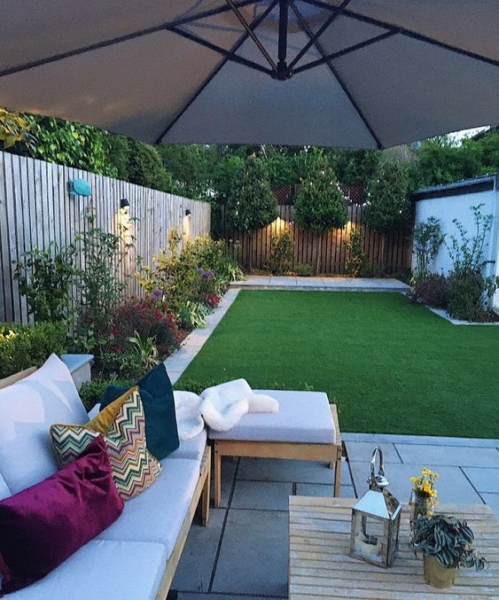 a modern garden with a green lawn, bright blooms lining it up, an outdoor sofa, an umbrella and some decor