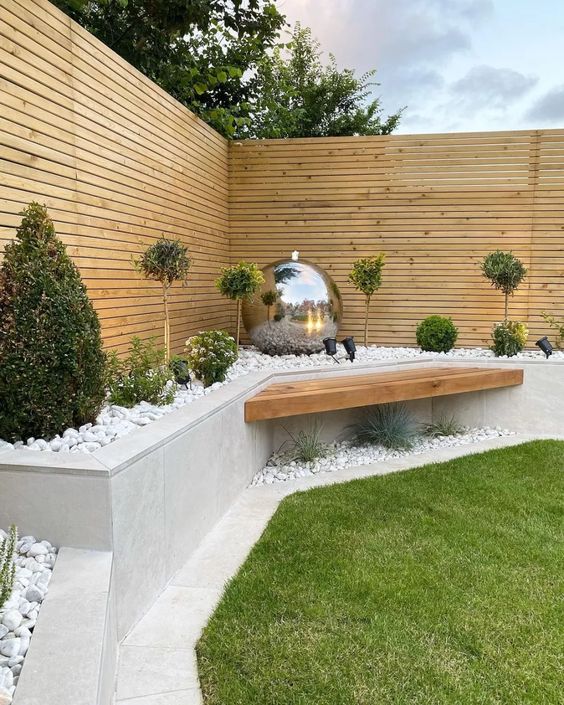 a modern garden with a green lawn, some plants in a raised garden bed, a built-in bench and a silver ball for decor