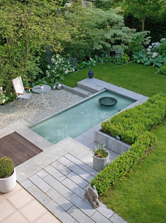 a modern garden with a green lawn, some shrubs, blooms and a water body plus some elegant garden furniture