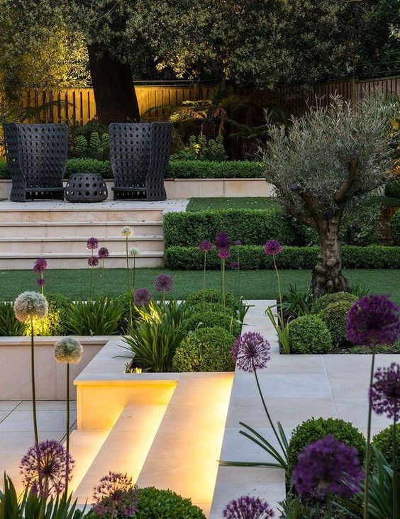 a modern garden with a green lawn, some shrubs, some blooms, a staircase with stunning modern chairs and built-in lights