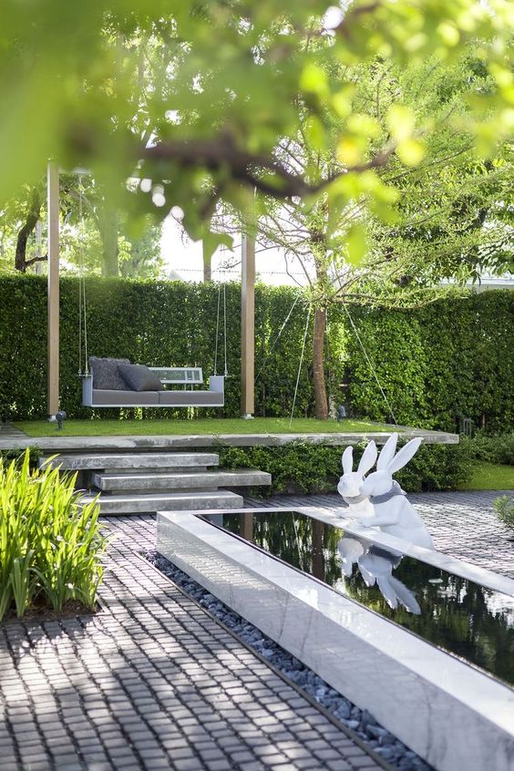 a modern garden with a water body, some grasses and greenery and swing sofa on a platform