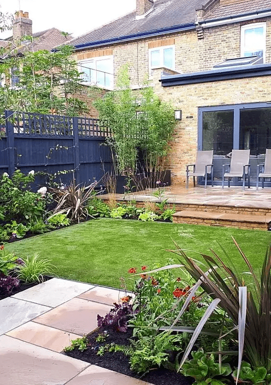 a modern garden with black privacy fences, a green lawn, some blooms and greenery, a deck with some chairs