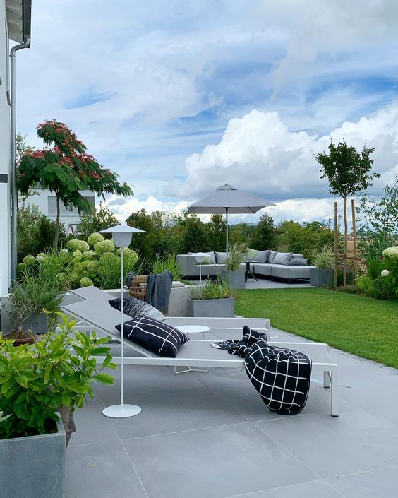 a modern garden with pavements, daybeds, floor lamps, a grey sofa, a green lawn and some trees and blooms in containers