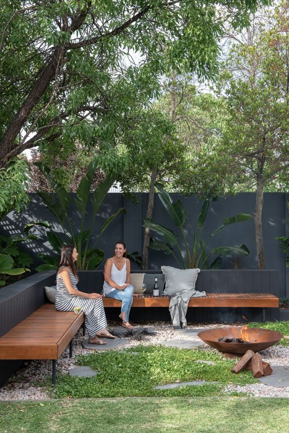 a modern garden with tropical plants, a stained bench, a fire pit and a green lawn is a cool and chic space