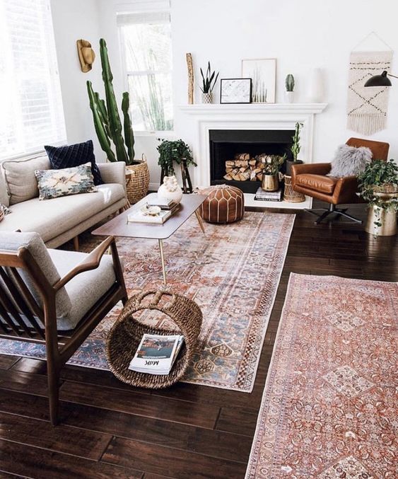 a modern living room with a fireplace, a neutral sofa and a cognac leather chair, potted plants, a coffee table, some boho decor on the mantel