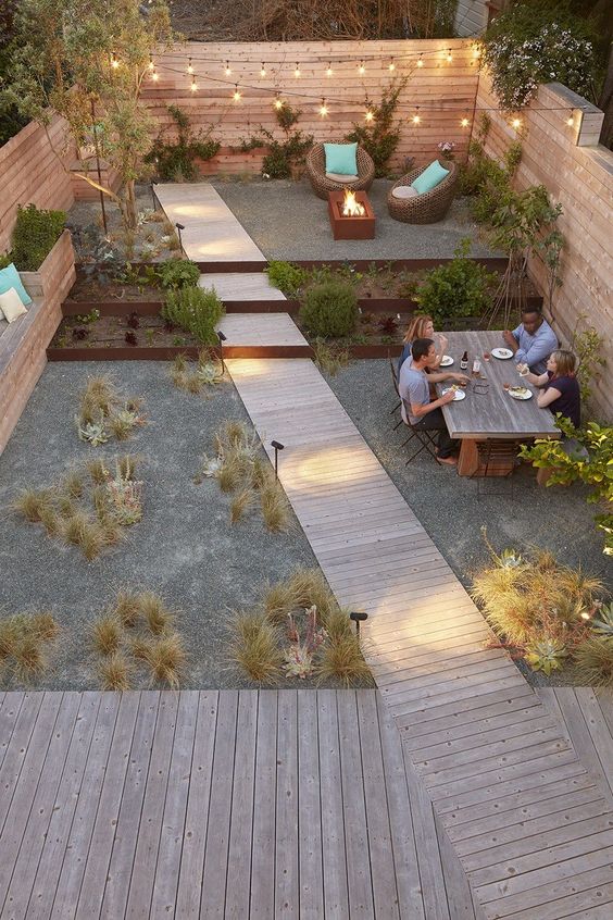 a modern multi-level outdoor space with a raised sitting zone with a fire pit, a dining space, some grasses and greenery