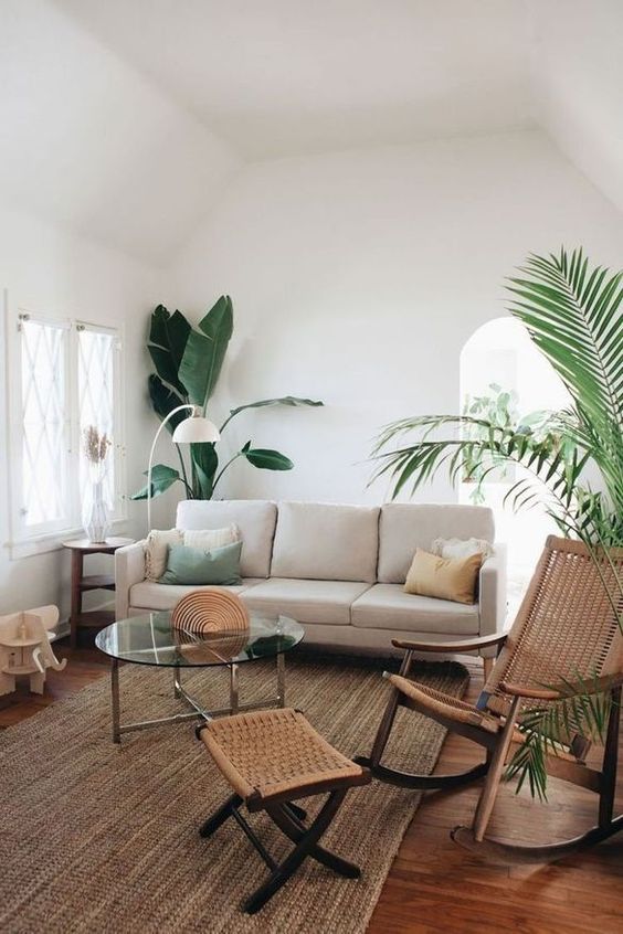 a modern neutral living room with a neutral sofa, a woven rocker and a footrest, a jute rug, a glass coffee table and potted plants is a chic space