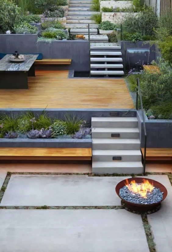 a modern nulti-level outdoor space with a dining space, a fountain, a bench with succulents and grasses, a fire pit