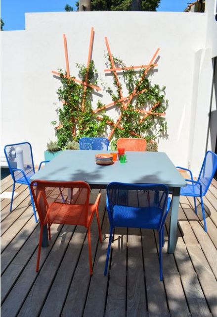 a modern outdoor dining space with a blue table, orange and blue chairs, an orange trellis with greenery on the wall
