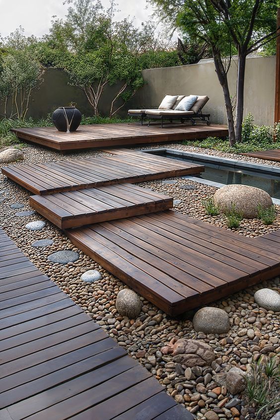 a modern outdoor space with a multi-level deck, a plunge pool, loungers, some greenery and trees around and some pebbles and rocks