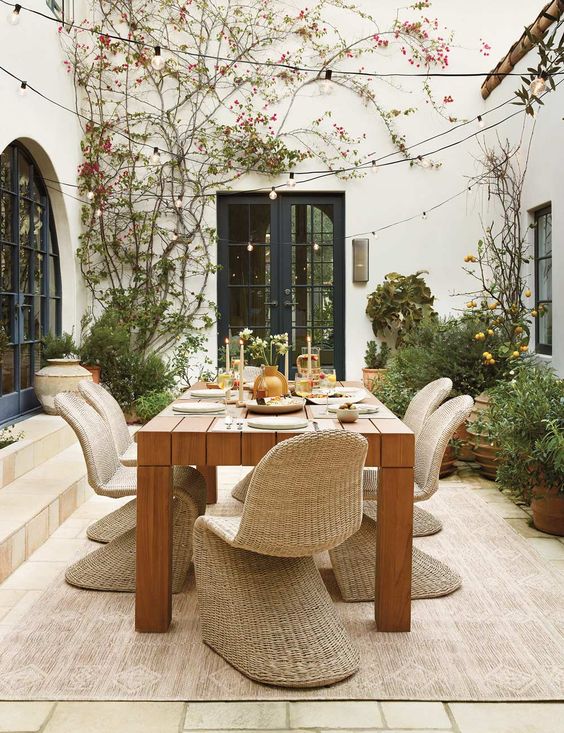 a modern rustic outdoor dining space with a stained table, chic wicker chairs and some greenery and lights around