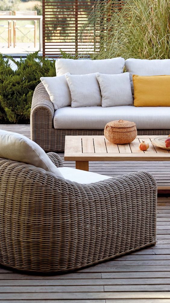 a modern rustic outdoor lounge with a wicker sofa and chair, a stained coffee table, some pillows and greenery around