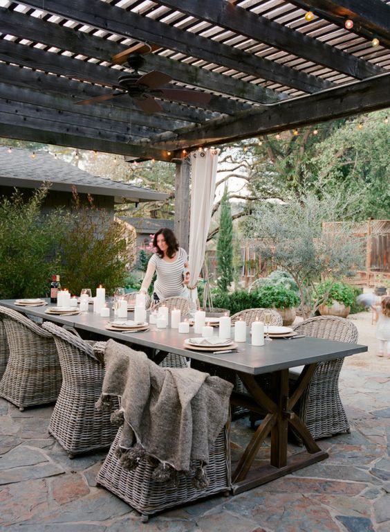 a modern rustic outdoor space with a concrete table, wicker chairs, greenery around and roof over the space