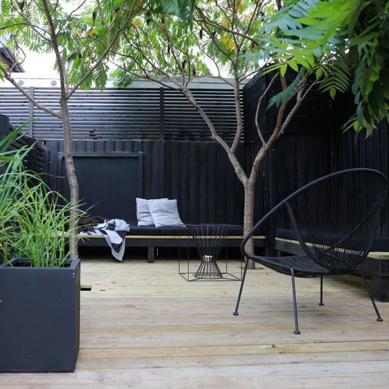 A modern terrace surrounded with a black fence, with trees and potted plants, a built in bench and some chairs