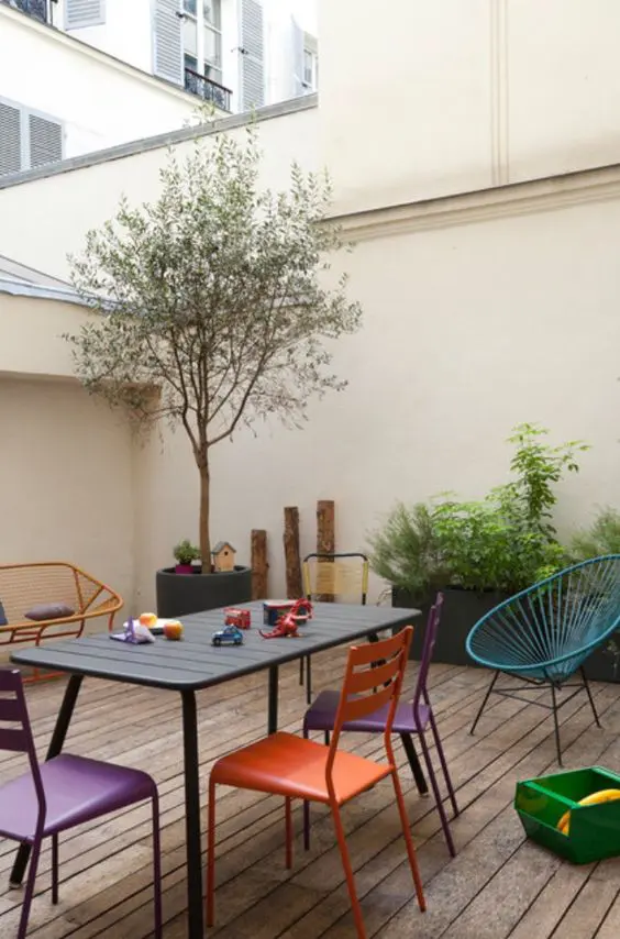 a modern terrace with a yellow loveseat, a grey table, purple and orange chairs, a blue chair, some greenery and a potted tree