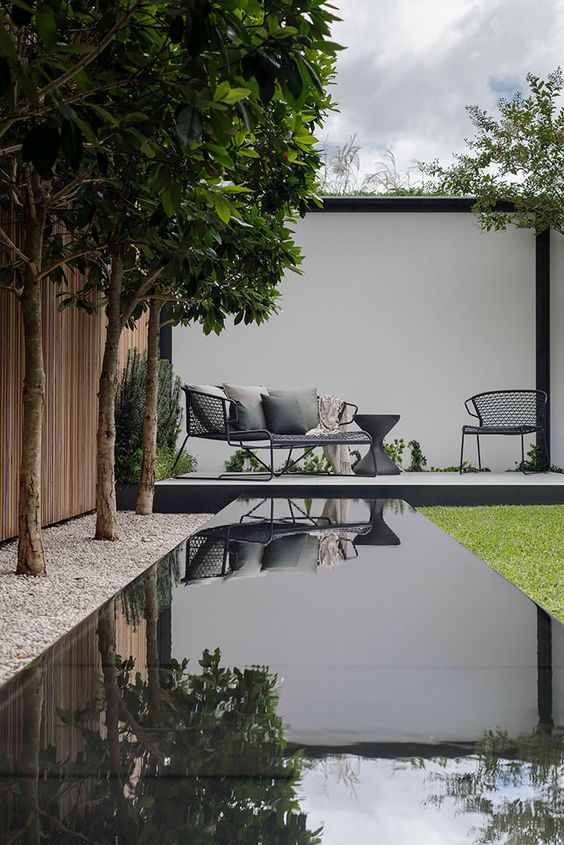 a modern to minimalist garden design with tall trees, a water body, a green lawn, a deck with black furniture