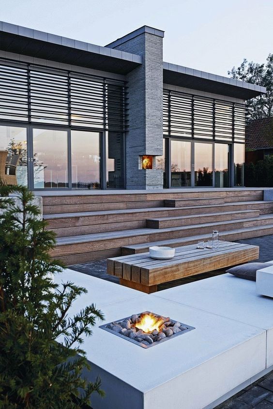 a multi-level deck, a sitting zone in the lower part, witha  fire pit, a wooden bench and some pillows and greenery around