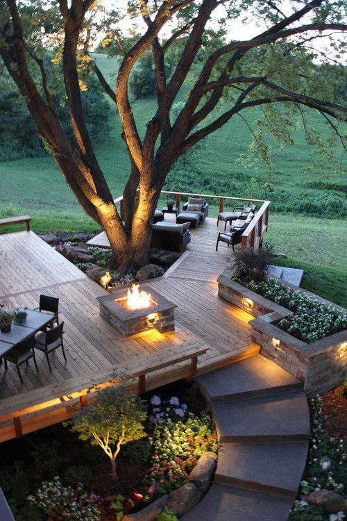 a multi-level outdoor space with a deck with an outdoor living room, a tree, blooms, a fire pit and a dining space