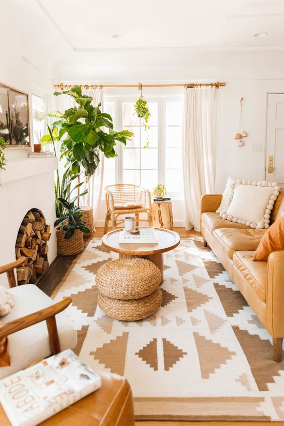 a neutral boho living room with a fireplace, a tan leather sofa, a boho rug, coffee tables, a rattan chair and some greenery