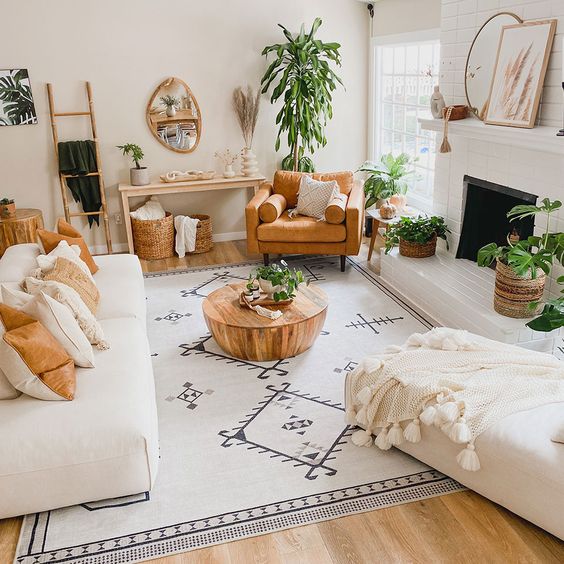 a neutral boho living room with a fireplace, creamy seatinf furniture, a boho rug, a coffee table, an amber leather chair, potted plants and a console with decor