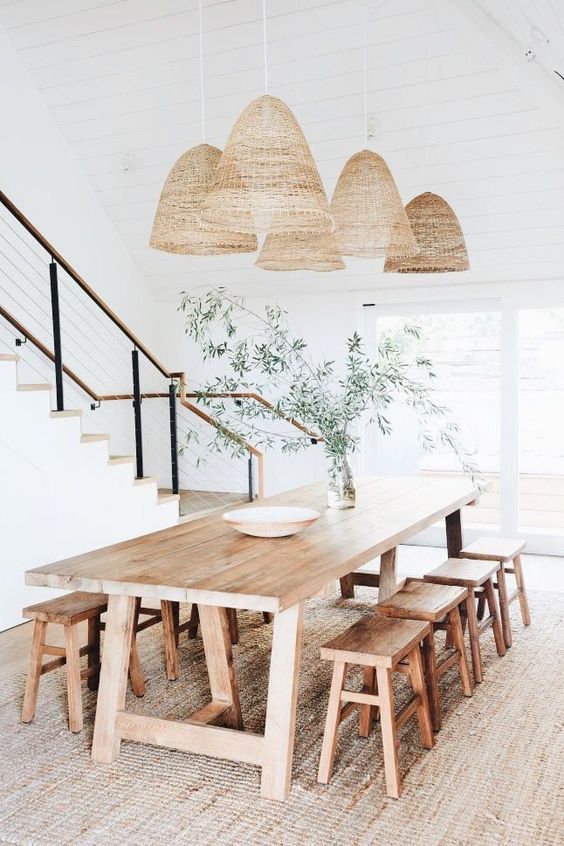 a neutral rustic dining room with a jute rug, a stained table and matching stools, woven pendant lamps and some greenery is airy and chic