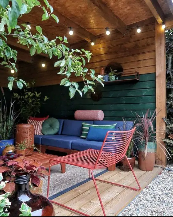 a pergola with a navy sofa, a pink metal chair, an orange table, colorful pillows, greenery and lights over the space