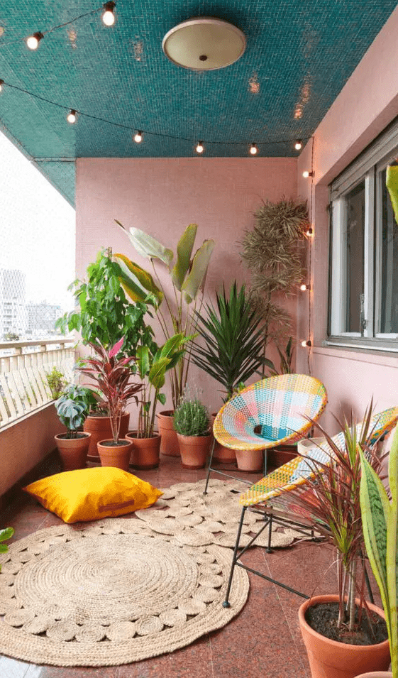 a pink balcony with colorful chairs, jute rugs, a yellow pillow and bold potted plants and lights