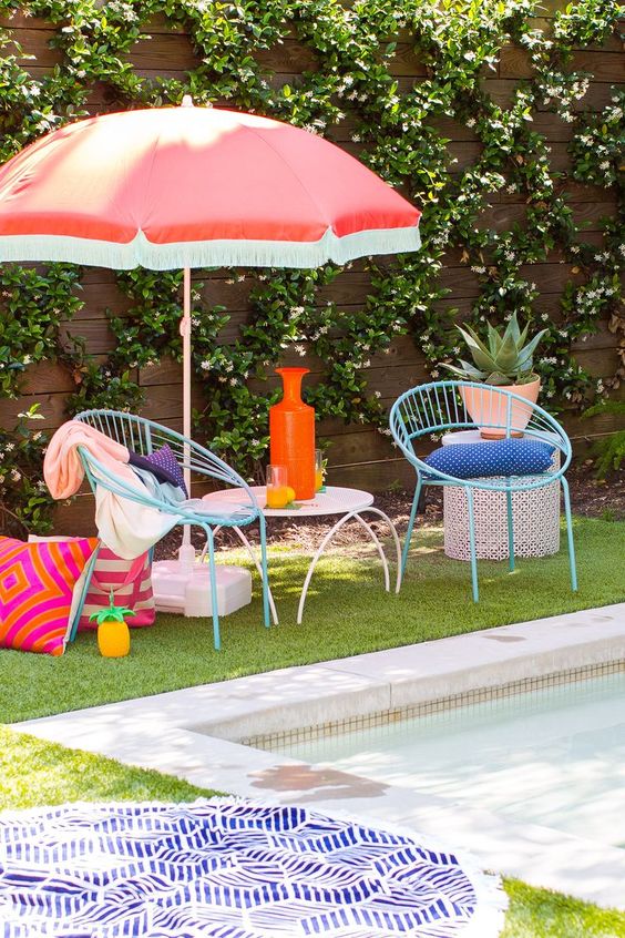 a poolside space with blue chairs, a side table, a red umbrella, some colorful pillows and decor
