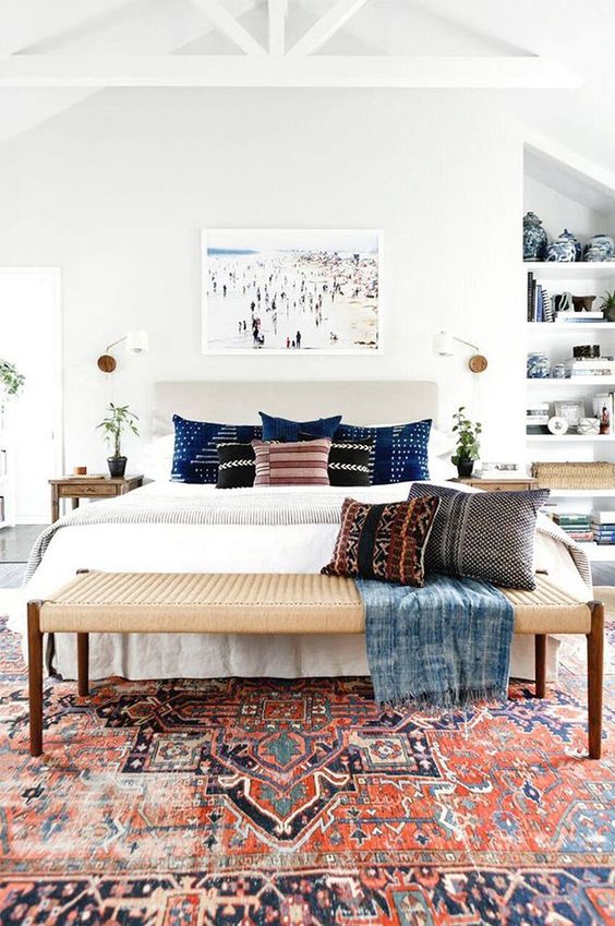 a pretty coastal bedroom with a bold boho rug, a neutral bed with printed pillows, a bench, built-in shelves and some potted plants