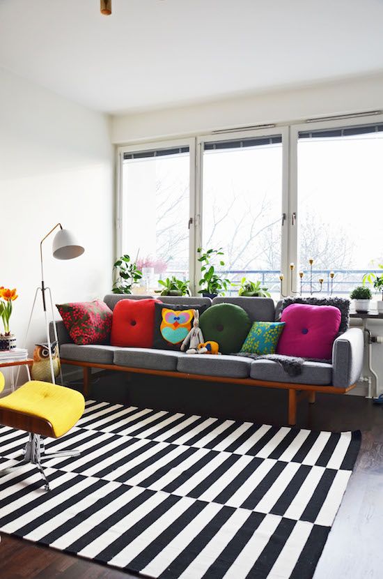 a pretty modern living room with a glazed wall, a grey sofa with colorful pillows, a striped rug, a yellow chair, a floor lamp and some greenery