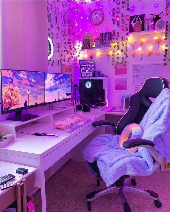 a purple gaming desk setup with a corner desk, purple lights, decor on the shelves, a chair with a blue blanket, a rainbow pillow and faux blooms