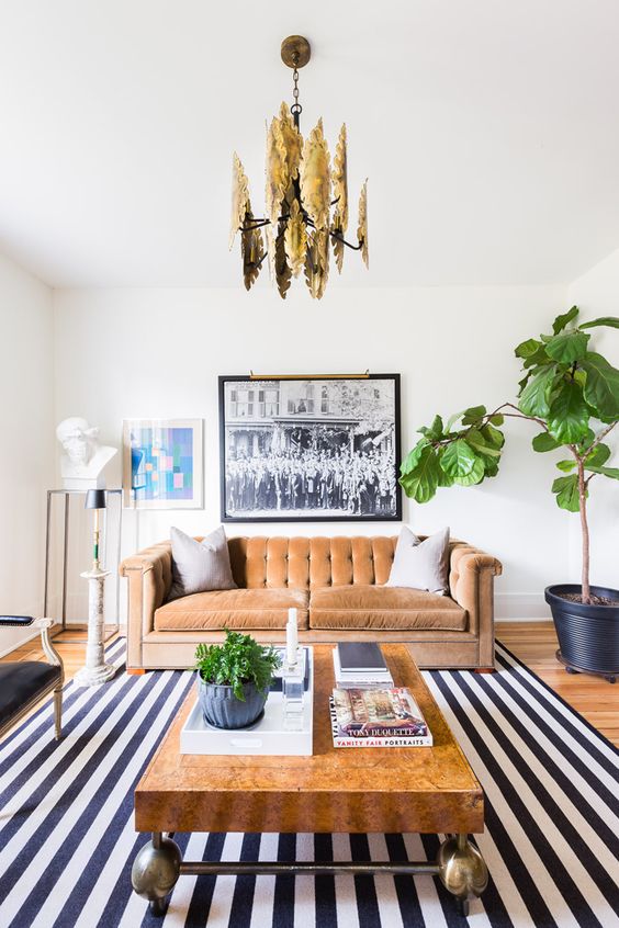 a quirky living room with a striped rug, a rust velvet sofa and a black loveseat, a coffee table, some art and a glam gold chandelier