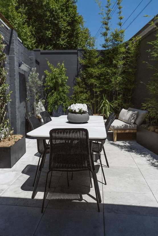 a small and stylish modern patio with a large table and black chairs, a daybed, greenery surrounding the space