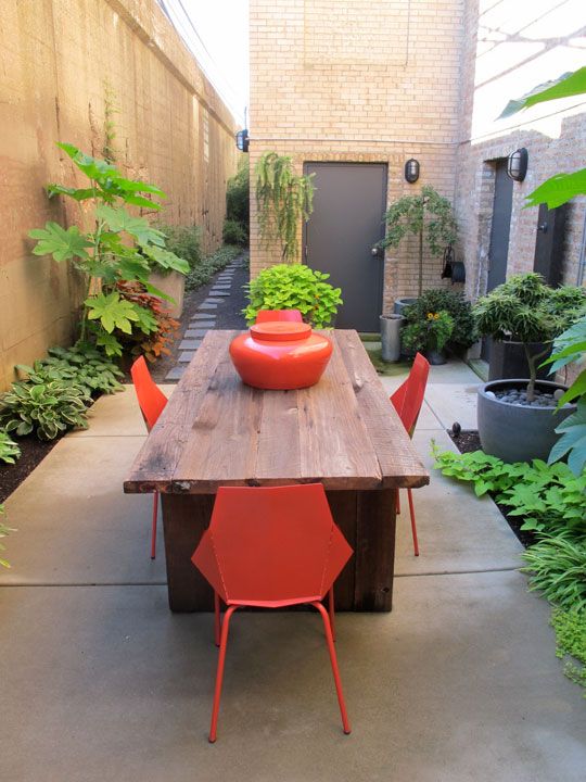 a small backyard with greenery, a stained table and fiery red plastic dinner chairs to add a bit of color to the space
