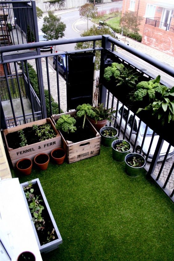 a small balcony with a green floor, boxes and planters on the floor and a box herb planter on the railing is a cool space with a garden