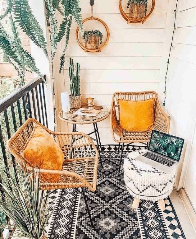 a small boho balcony with a black and white rug, rattan chairs with yellow pillows, a black and white pouf, potted plants and hanging planters