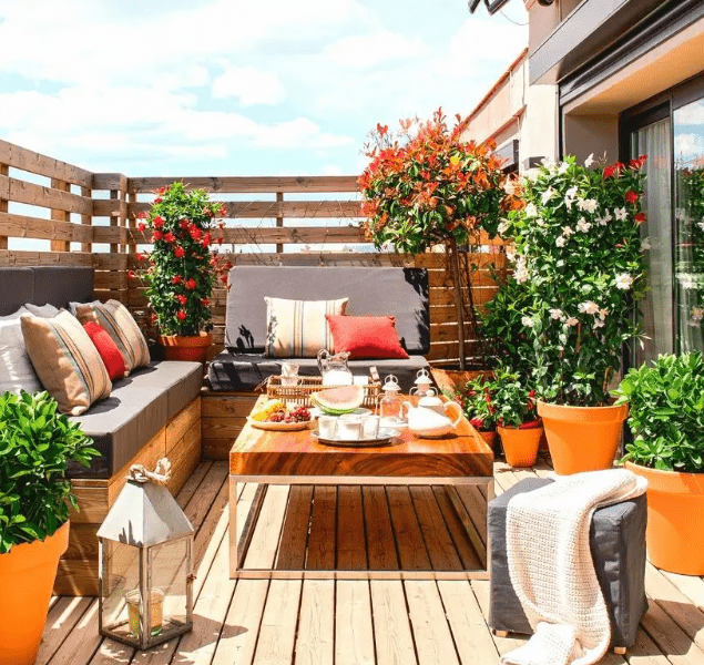 a small colorful terrace with a built-in corner seat, a table, potted greenery and blooms and a candle lantern