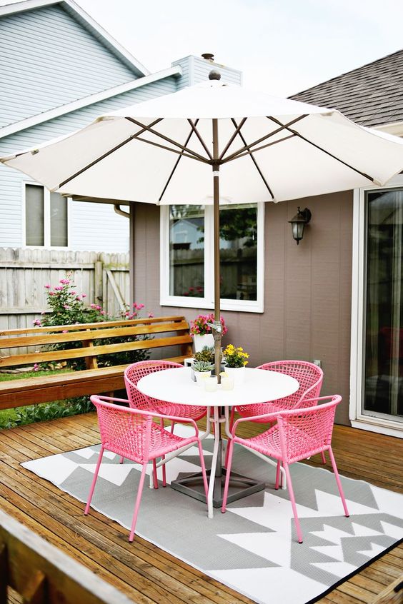 a small deck with a stained bench, an umbrella with a table, pink wicker chairs and a rug is a cool space to have a meal