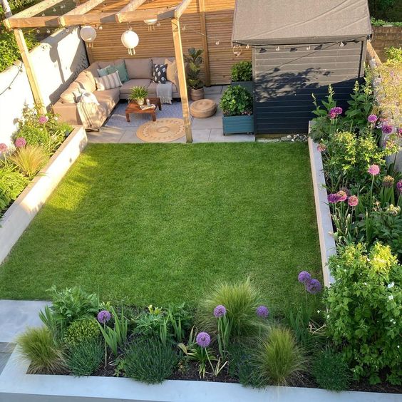 a small modern garden with a green lawn, raised beds with blooms and greenery, a small sitting space and lights is cool