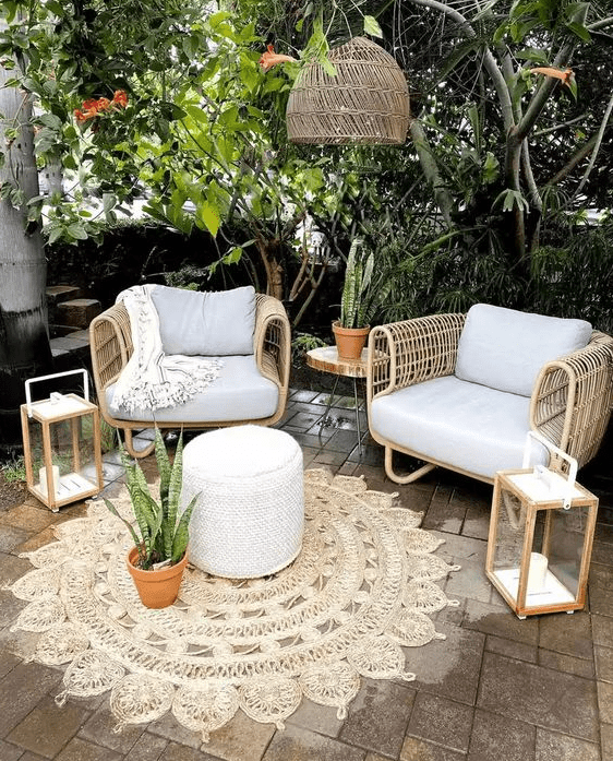 a small patio with wicker furniture, a pouf, some candle lanterns, a woven pendant lamp and greenery around